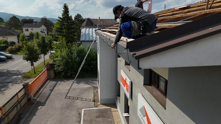Ein Handwerker kniet auf einem Hausdach und führt eine Dachrinne Montage durch, während er ein Kabel an der Dachkante befestigt.