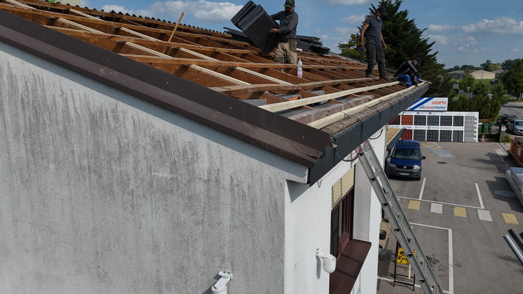 Mehrere Arbeiter führen eine Dachrinne Montage und Dacharbeiten auf einem freigelegten Dachstuhl durch; einer trägt Dachplatten, während andere entlang der Dachkante arbeiten. Eine Leiter lehnt an der Fassade, unten ist ein Parkplatz mit einem Fahrzeug zu sehen.